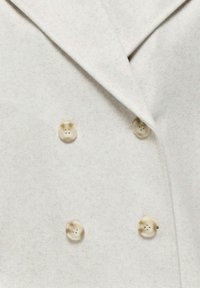 Manteau croisé beige clair avec quatre grands boutons en écaille de tortue et un large col à revers cranté.