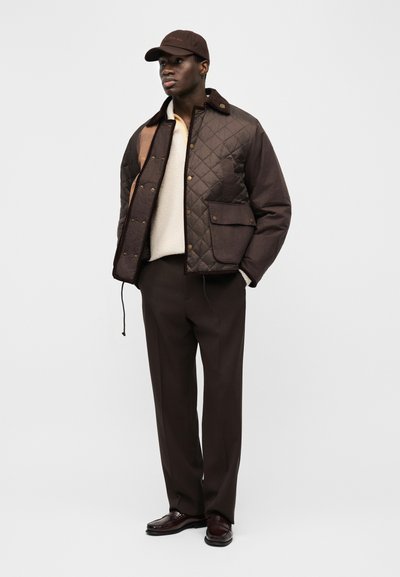 Moški, oblečen v rjavo prešito jakno, krem pulover, rjave hlače, ujemajočo rjavo kapo in temno rjave čevlje, stoji pred enobarvnim ozadjem.