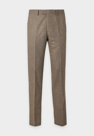 Homme portant une chemise beige clair à manches longues boutonnée, un pantalon vert olive et des chaussures habillées marron foncé, debout devant un fond uni.