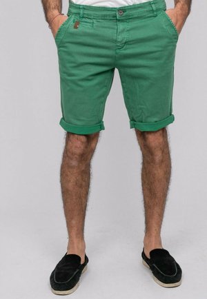 Homme portant un short vert retroussé, des chaussures noires slip-on, les mains dans les poches, debout devant un fond uni.