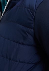 Navy modrý prošívaný vest, s horizontálně prošívanými sekcemi, kontrastním texturovaným límcem a předním zipem s bílými zuby.