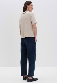 Donna in piedi di profilo, che indossa un top a manica corta a maglia beige, jeans a gamba larga blu scuro e mocassini neri su uno sfondo bianco.