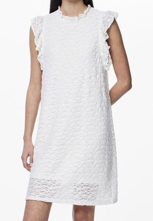 Vrouw draagt een mouwloze witte jurk met een gestructureerd patroon en gerimpelde armsgaten, staand met één arm achter haar rug tegen een witte achtergrond.