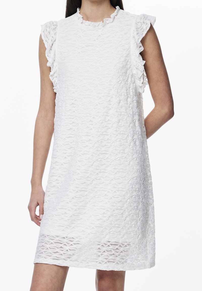 Femme portant une robe blanche texturée sans manches avec des emmanchures volantes, debout avec un bras derrière le dos sur un fond blanc.