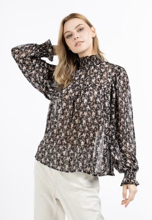 Frau mit blondem Haar, die eine schwarze transparente Bluse mit kleinem weißem Blumenmuster und cremefarbene Hose trägt, posiert mit einer Hand hinter dem Kopf.