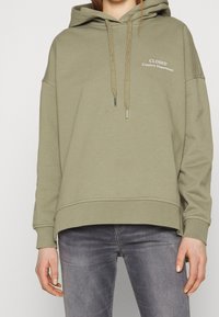 Sweat à capuche kaki avec cordon de serrage et poche avant. Inscription "CLOSED Creative Department" en blanc sur le côté gauche de la poitrine. Tissu doux, coupe décontractée.