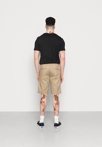 Cămașă neagră cu mânecă scurtă și decolteu rotund, asortată cu pantaloni scurți bej până la genunchi. Adidași negri și șosete albe până la gleznă vizibile.