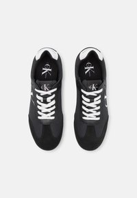 Černé tenisky vyrobené z látky a kožešiny s bílými šněrovadly, s logem na boku a polstrovaným límcem pro pohodlí.