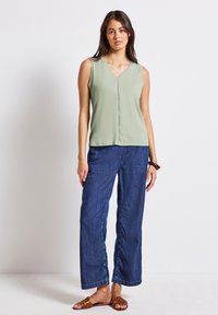 Top verde senza maniche con scollatura a V e dettaglio cucitura al centro, abbinato a pantaloni in denim blu scuro a vestibilità ampia e sandali piatti marroni.