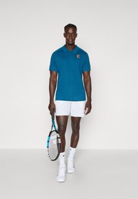 Tyrkysové polo tričko s golierom a logom, biele šortky, tenisová raketa v ruke, biele tenisky, umiestnené proti minimalistickému pozadiu.