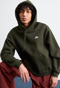Dark green hoodie with a front pocket, drawstring hood, and white Nike SB logo on the left chest; worn with maroon pants.