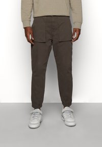 Brązowe spodnie cargo z elastycznymi mankietami, przednimi kieszeniami i luźnym krojem, połączone z białymi trampkami. Tkanina wydaje się miękka i wygodna.