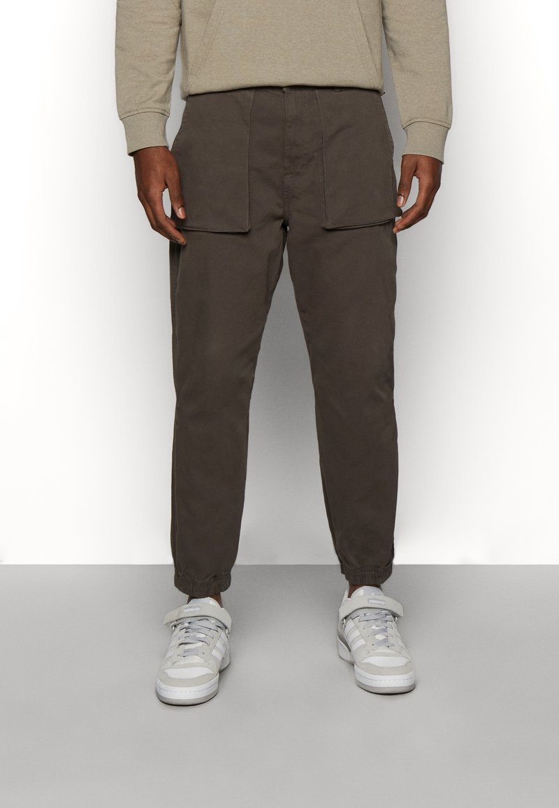 Brązowe spodnie cargo z elastycznymi mankietami, przednimi kieszeniami i luźnym krojem, połączone z białymi trampkami. Tkanina wydaje się miękka i wygodna.