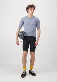 Maillot de cyclisme gris à manches courtes et fermeture éclair, associé à un cuissard de cyclisme noir et des chaussettes jaunes ; tenant un casque gris.