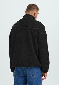 Chaqueta de forro polar negra con un collar alto, superficie texturizada, corte relajado y mangas largas, llevada sobre unos jeans de mezclilla azules.