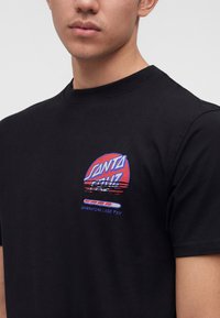 Νέος άνδρας φορώντας μαύρο T-shirt με παραμορφωμένο κόκκινο και μπλε λογότυπο "Santa Cruz" και εικονίδιο μπάρας φόρτωσης στο αριστερό στήθος.