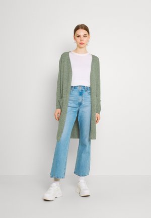 Lichtgroene gebreide cardigan met open voorkant, lange mouwen en een losse fit, gedragen over een wit T-shirt en hooggewaiste blauwe spijkerbroek.