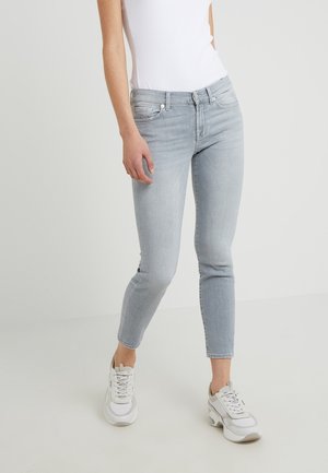 Persona con jeans ajustados grises claros, zapatillas blancas y una blusa blanca sin mangas, de pie contra un fondo plano.