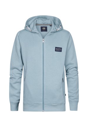 Garçon portant un sweat à capuche zippé bleu clair, un jean gris foncé et des baskets blanches, debout devant un fond blanc uni.