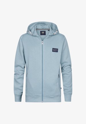 Garçon portant un sweat à capuche zippé bleu clair, un jean gris foncé et des baskets blanches, debout devant un fond blanc uni.
