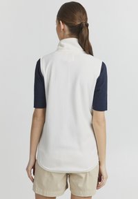 Weißer Fleece-Weste mit hohem Kragen, ärmellosem Design und abgerundetem Saum, getragen über einem dunklen gerippten Shirt und kombiniert mit hellbeigen Shorts.