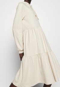 Robe à capuche couleur crème avec des manches longues, dotée d'une jupe à volants et de détails froncés. Fabriquée en tissu doux et texturé.