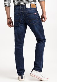 Ciemnoniebieskie dżinsy o slim fit, z widocznym przeszyciem, dwiema tylnymi kieszeniami i skórzaną naszywką w pasie. Noszone z białymi trampkami.