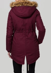 Burgundy parka-jacka med huva i fuskpäls, åtdragen midja, elastiska ärmmuddar och splittad nederkant. Tillverkad i textil med struktur för värme.