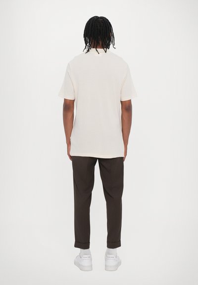 T-shirt beige clair à manches courtes et coupe décontractée, associé à un pantalon marron retroussé et des baskets blanches.
