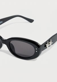 Lunettes de soleil noires avec montures ovales, verres teintés foncés et accents en forme d'étoile argentée sur les branches. Fini lisse et brillant, détails minimalistes.