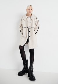 Faux fur frakke i beige med en stor krave, sorte knapper og kontrasterende sorte sømme. Båret over et sort sæt tøj og parret med kraftige sorte støvler.