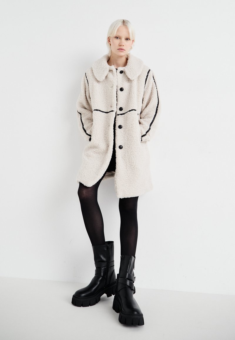 Beige Kunstpelzjacke mit großem Kragen, schwarzen Knöpfen und kontrastierenden schwarzen Nähten. Getragen über einem schwarzen Outfit und kombiniert mit klobigen schwarzen Stiefeln.