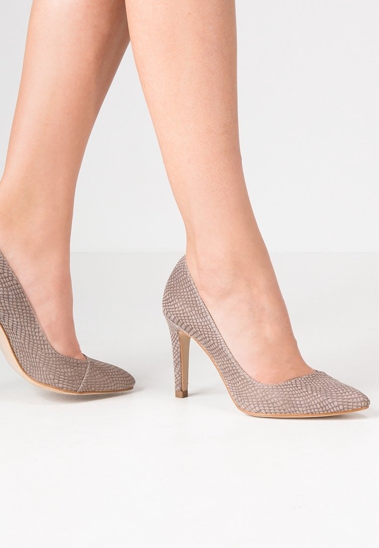 Jambes féminines portant des escarpins à talons hauts gris texturés, debout sur une surface blanche avec un fond blanc uni.