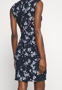 Ärmlös, knälång klänning i marinblått tyg med ett blommönster av lila och vita blommor. Har en figursydd midja och sömdetaljer bak.