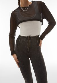 Crop top a maniche lunghe in rete nera sovrapposto a una maglietta aderente bianca, abbinato a pantaloni in denim grigio scuro a vita alta con chiusura zip.