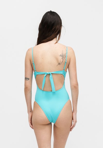 Γυναίκα με μακριά σκούρα μαλλιά που φοράει ολόσωμο μαγιό ανοιχτού μπλε χρώματος με λεπτές τιράντες και ανοιχτή πλάτη δεμένη με φιόγκο.