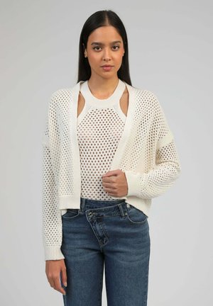 Femme portant un cardigan blanc en maille ouverte sur un haut sans manches assorti, associé à un jean bleu taille haute, debout devant un fond uni.