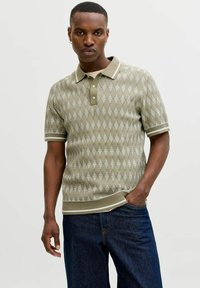 Junger Mann, der ein kurzärmeliges, olivgrünes, gemustertes Poloshirt aus Strick mit weißem Besatz, dunkle Jeans und kleine Creolen trägt und mit einer Hand in der Tasche steht.