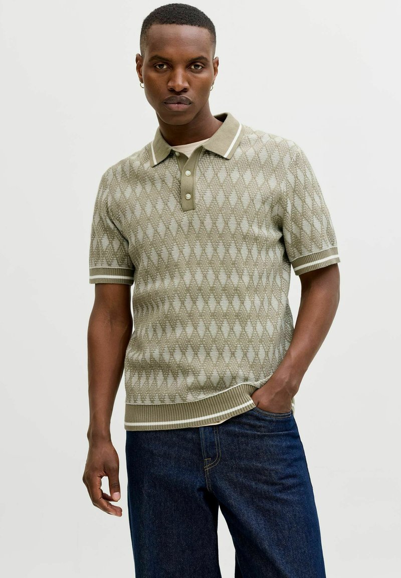 Junger Mann, der ein kurzärmeliges, olivgrünes, gemustertes Poloshirt aus Strick mit weißem Besatz, dunkle Jeans und kleine Creolen trägt und mit einer Hand in der Tasche steht.