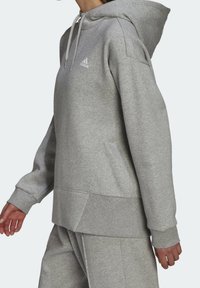 Personne portant un sweat à capuche Adidas gris clair et un pantalon de survêtement assorti, montrée du cou jusqu'au milieu des cuisses, les mains détendues le long du corps.