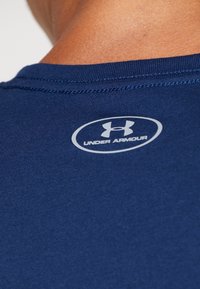 Granatowa koszulka sportowa o gładkiej fakturze, dopasowanym kroju i białym logo Under Armour z tyłu, blisko dekoltu.