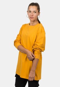 Langärmeliges, oversized T-Shirt aus leuchtend gelbem Baumwollstoff. Mit Rundhalsausschnitt und hochgerollten Ärmeln. Glatte Textur mit minimalen Nähten.