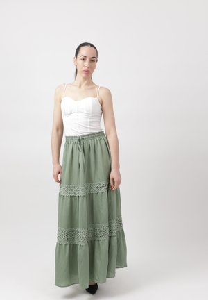 Vrouw staand met een witte mouwloze top en een lange groene rok met kanten details tegen een effen lichte achtergrond.