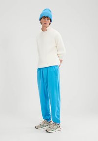 Krēmkrāsas ribota džemperis, spilgti zili sporta bikses ar kabatām un balti kedu ar zaļiem akcentiem. Uz galvas zila adīta cepure.