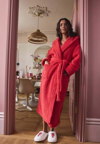 Red, fluffy bathrobe with a tie waist and two pockets. Worn with white slippers featuring a lobster design. Wooden floor and pink walls in background.