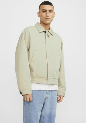 Beige cropped jakke med bred krave, to forlommer og knapdetaljer. Bæres over en grå skjorte og parret med blå denim jeans.