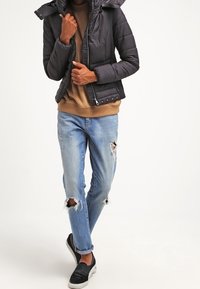 Kort svart vadderad jacka med huva, över en beige sweatshirt. Ljusblå slitna jeans och svarta slip-on skor. Avslappnad passform.