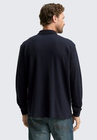Marineblaues Langarm-Polo-Shirt aus strukturiertem Stoff, mit Kragen und seitlichen Schlitzen, kombiniert mit hellblauen Jeans.