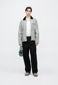 Jachetă gri cu guler negru, bluză crem, pantaloni negri cu picioare largi și geantă verde ținută în mână. Modelul poartă pantofi gri slip-on.