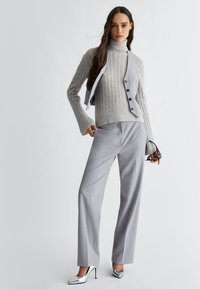 Maglione grigio chiaro a trecce con maniche ampie, indossato sopra un gilet grigio con bottoni. Abbinato a pantaloni grigi chiari e tacchi argentati lucidi.